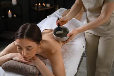 Photo of Esthetician applying cosmetic product for body wraps treatment onto woman's back in spa salon, closeup