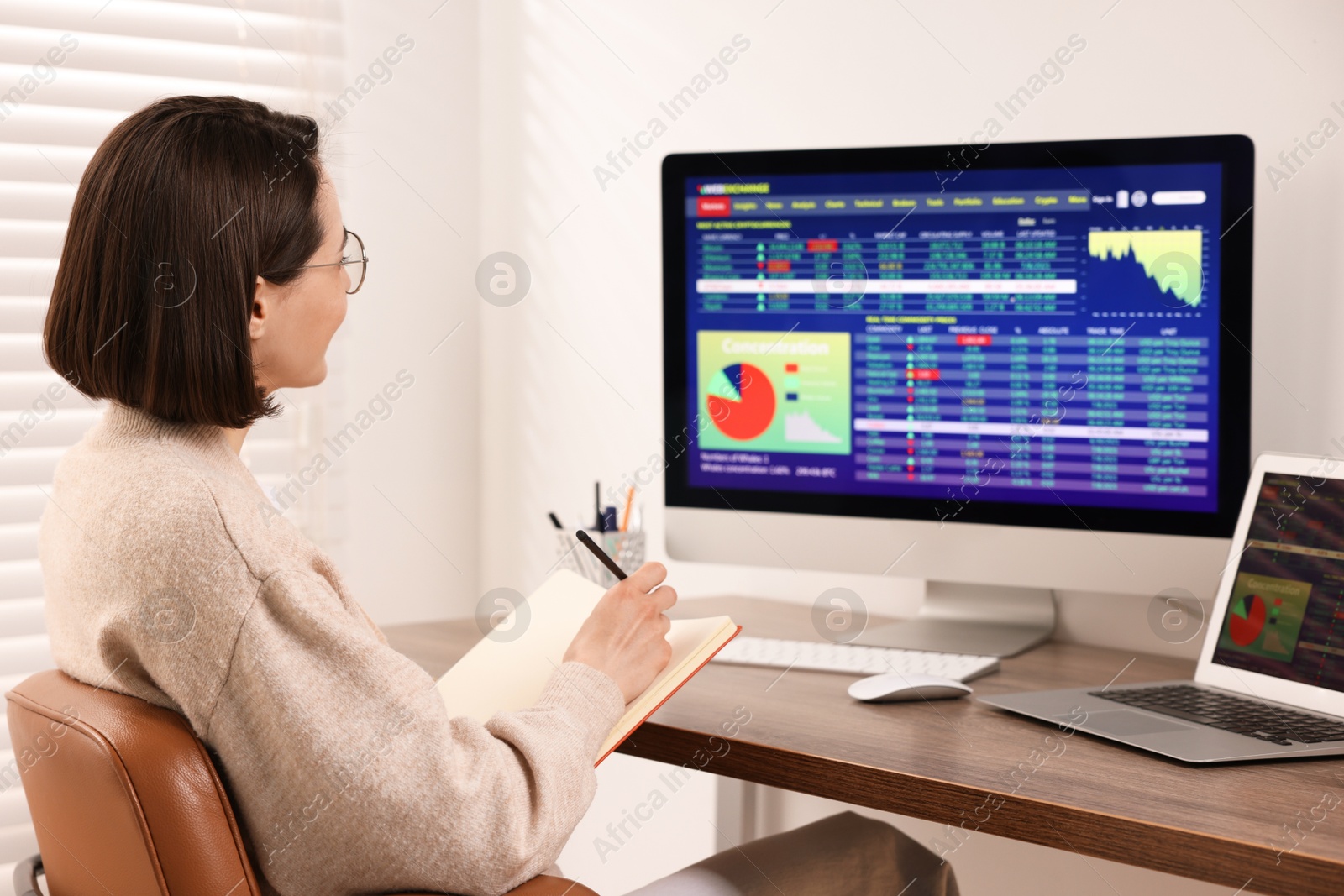 Photo of Stock exchange. Woman taking notes while analysing financial market on computer at wooden table indoors