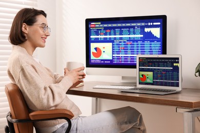 Photo of Stock exchange. Woman with cup of drink analysing financial market at wooden table indoors