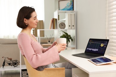 Stock exchange. Woman with cup of drink analysing financial market on laptop at white table indoors