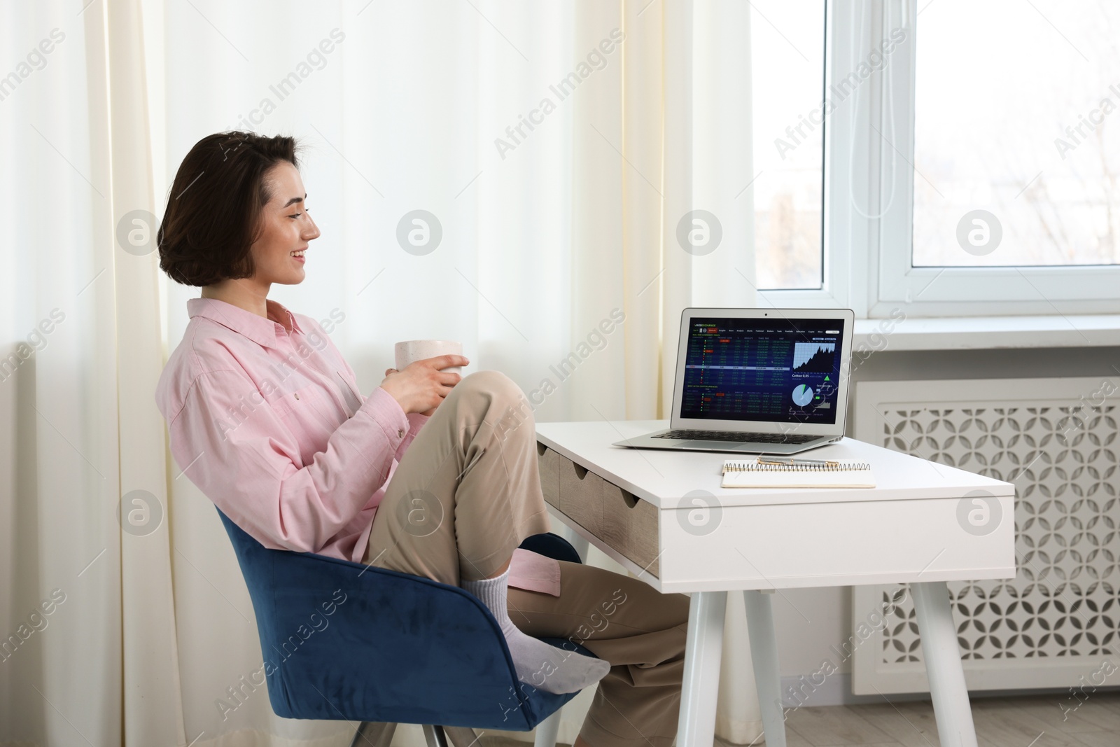 Photo of Stock exchange. Woman with cup of drink analysing financial market on laptop at white table indoors