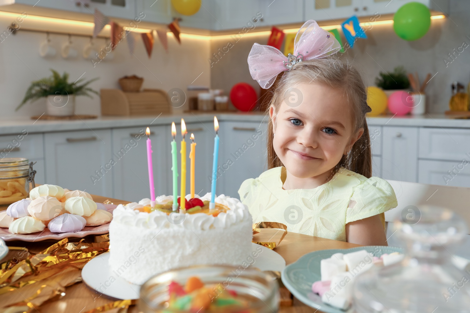 Photo of Cute little girl at table with birthday cake and different treats indoors. Surprise party