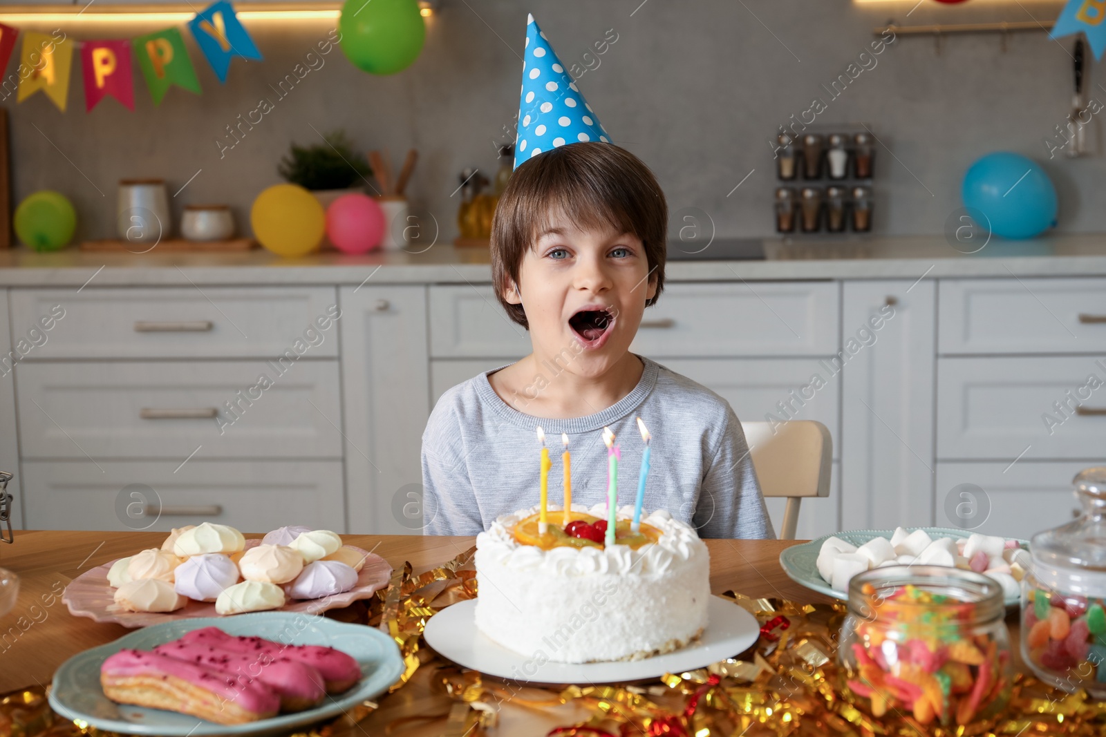 Photo of Cute little boy in conical paper hat at table with birthday cake and different treats indoors. Surprise party