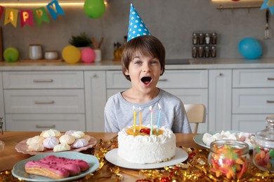 Photo of Cute little boy in conical paper hat at table with birthday cake and different treats indoors. Surprise party