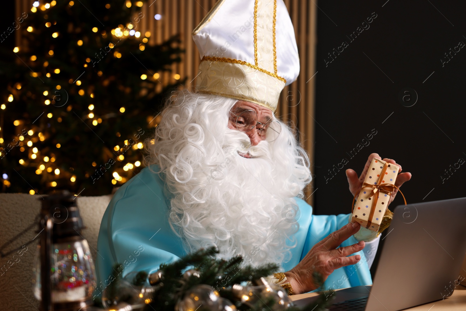 Photo of Saint Nicholas with gift and laptop in room decorated for Christmas