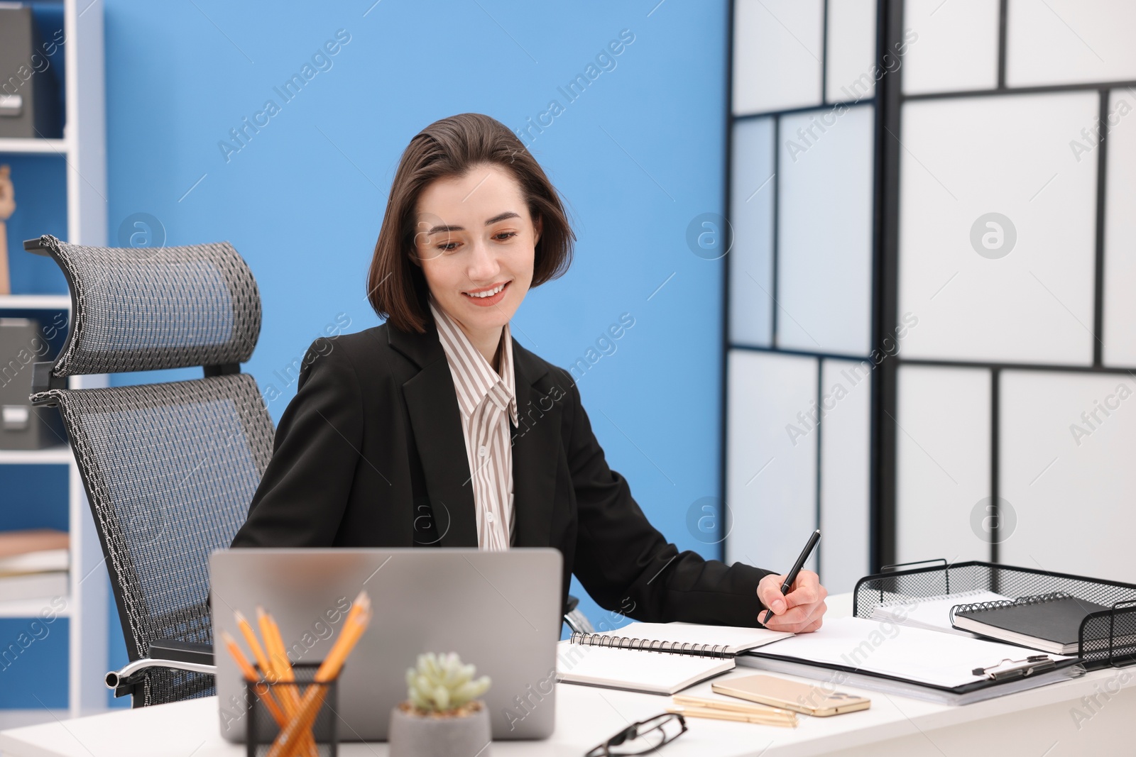 Photo of Smiling secretary taking notes while working on laptop at table in office