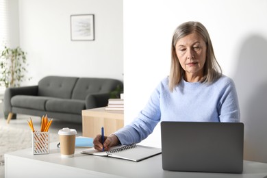 Photo of Woman learning online using laptop and taking notes at table indoors. Self-study