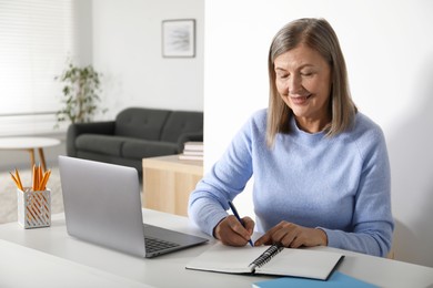 Photo of Smiling woman learning online using laptop and taking notes at table indoors. Self-study