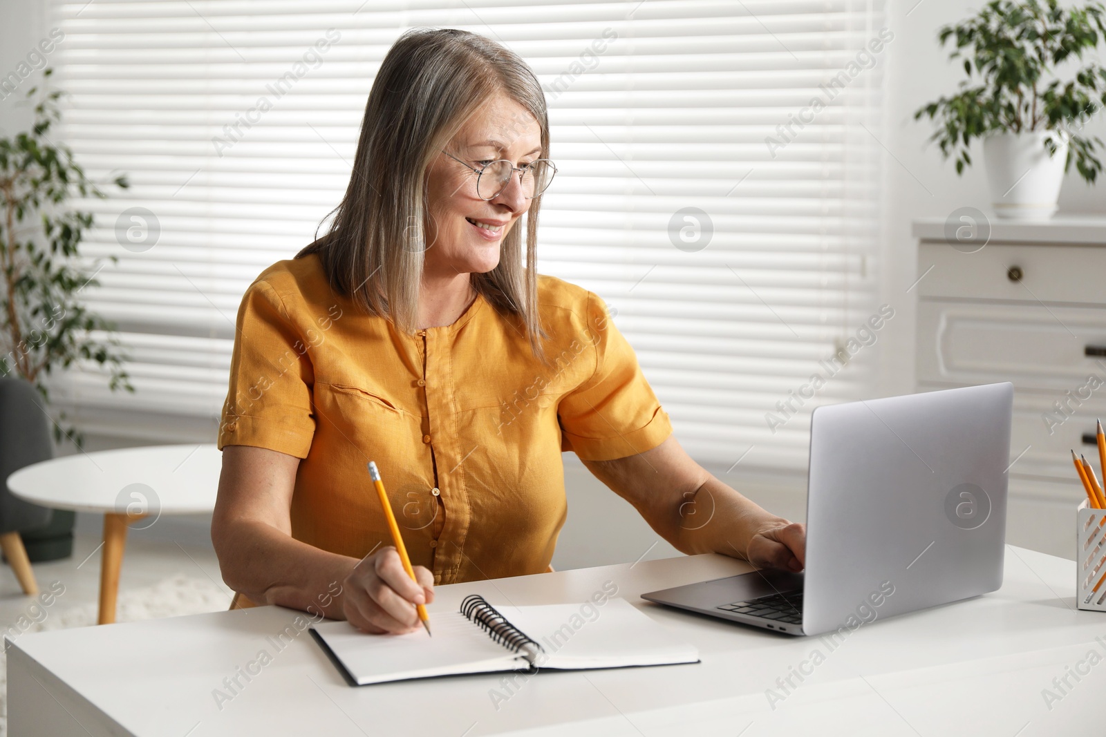 Photo of Smiling woman learning online using laptop and taking notes at table indoors. Self-study