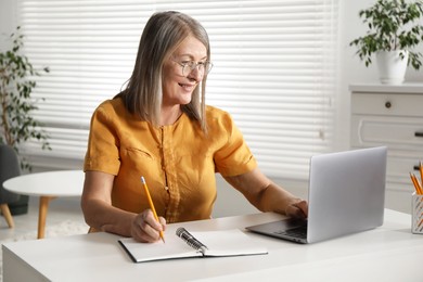 Photo of Smiling woman learning online using laptop and taking notes at table indoors. Self-study