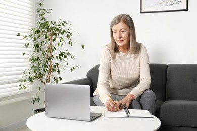 Photo of Woman learning online using laptop and taking notes at table indoors. Self-study