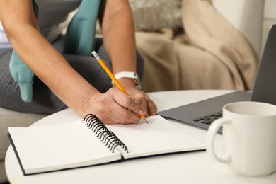 Photo of Woman learning online using laptop and taking notes at white table indoors, closeup. Self-study