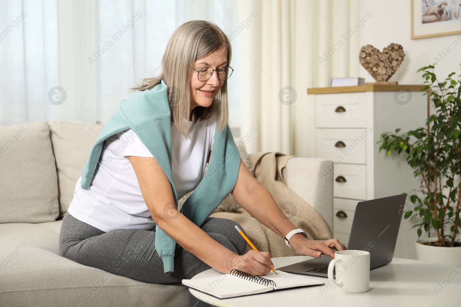 Photo of Smiling woman learning online using laptop and taking notes at table indoors. Self-study