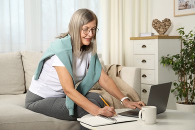 Smiling woman learning online using laptop and taking notes at table indoors. Self-study