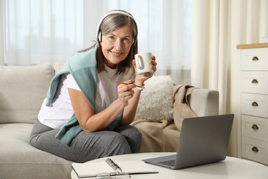 Smiling woman with cup learning online using laptop at table indoors. Self-study