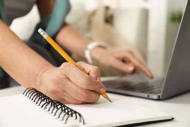 Photo of Woman learning online using laptop and taking notes at white table indoors, closeup. Self-study