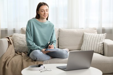 Photo of Smiling woman taking notes during online lesson on sofa indoors. Self-study