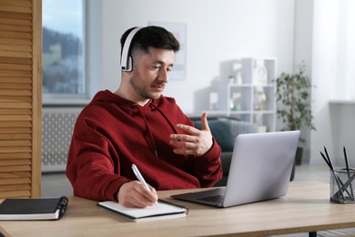 Man taking notes during online lesson with teacher at table indoors