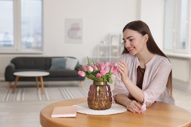 Photo of Smiling woman with bouquet of pink tulips at table indoors. Space for text