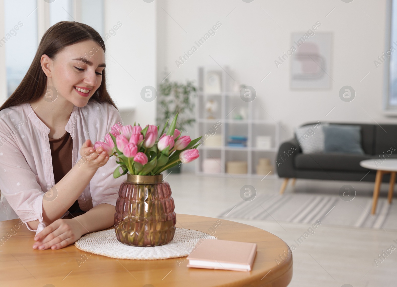 Photo of Smiling woman with bouquet of pink tulips at table indoors. Space for text