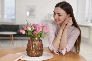 Photo of Smiling woman with bouquet of pink tulips at table indoors