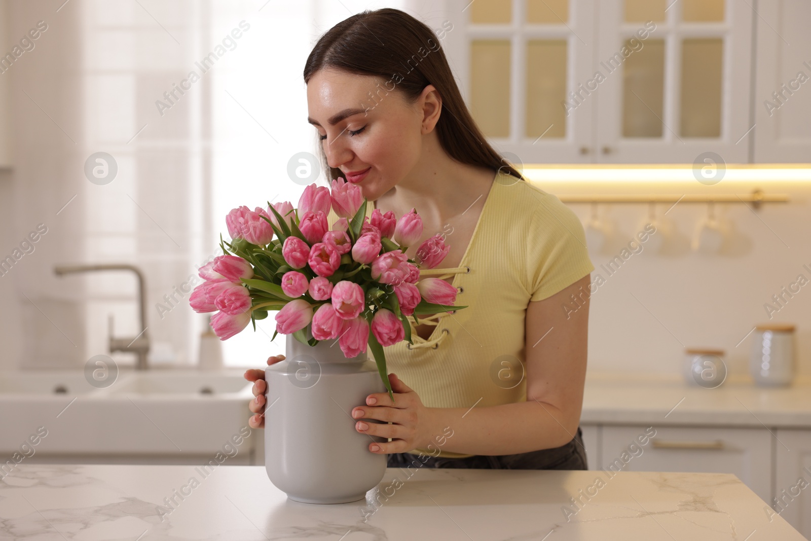 Photo of Woman sniffing bouquet of pink tulips at table in kitchen