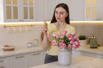 Woman putting tulip into vase with bouquet at table in kitchen