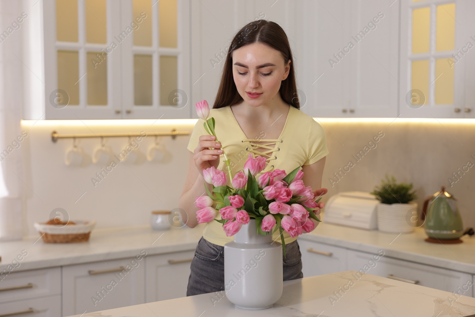 Photo of Woman putting tulip into vase with bouquet at table in kitchen