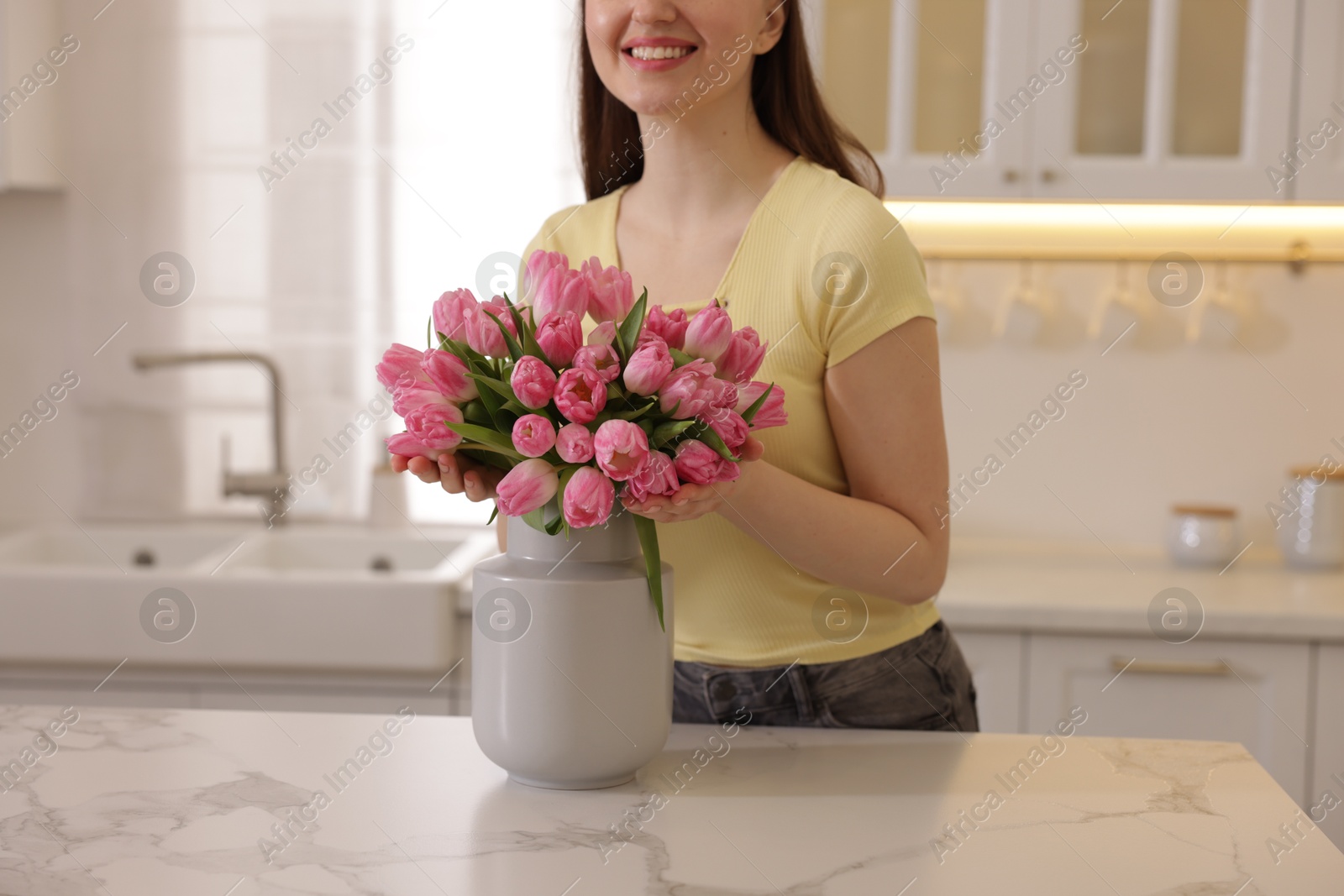 Photo of Smiling woman with bouquet of pink tulips at table in kitchen, closeup