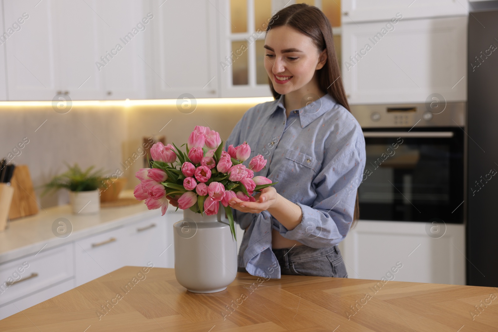 Photo of Smiling woman with bouquet of pink tulips at table in kitchen