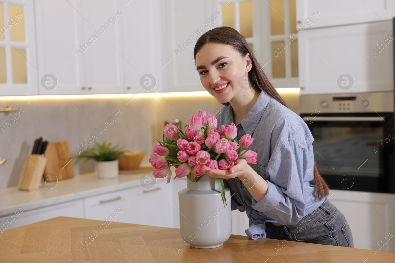 Photo of Smiling woman with bouquet of pink tulips at table in kitchen