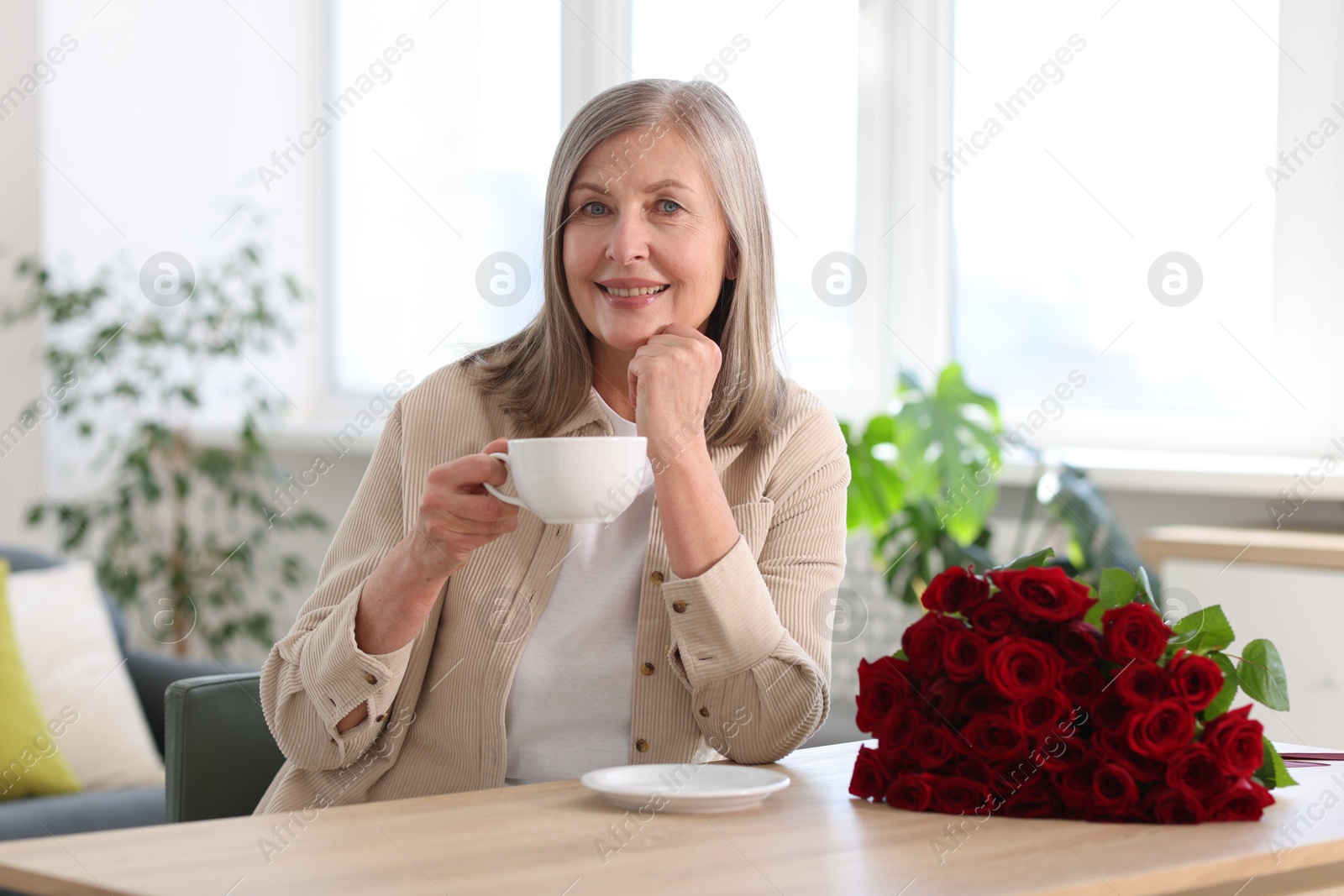 Photo of Smiling woman with bouquet of roses drinking at table indoors