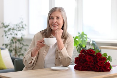 Smiling woman with bouquet of roses drinking at table indoors