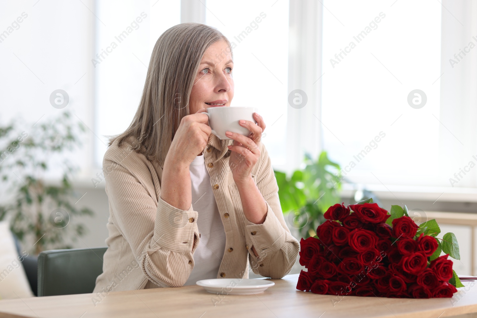 Photo of Smiling woman with bouquet of roses drinking at table indoors