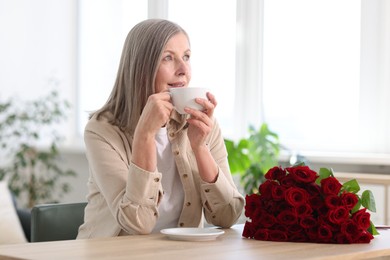 Smiling woman with bouquet of roses drinking at table indoors