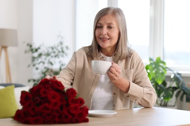 Smiling woman with bouquet of roses drinking at table indoors