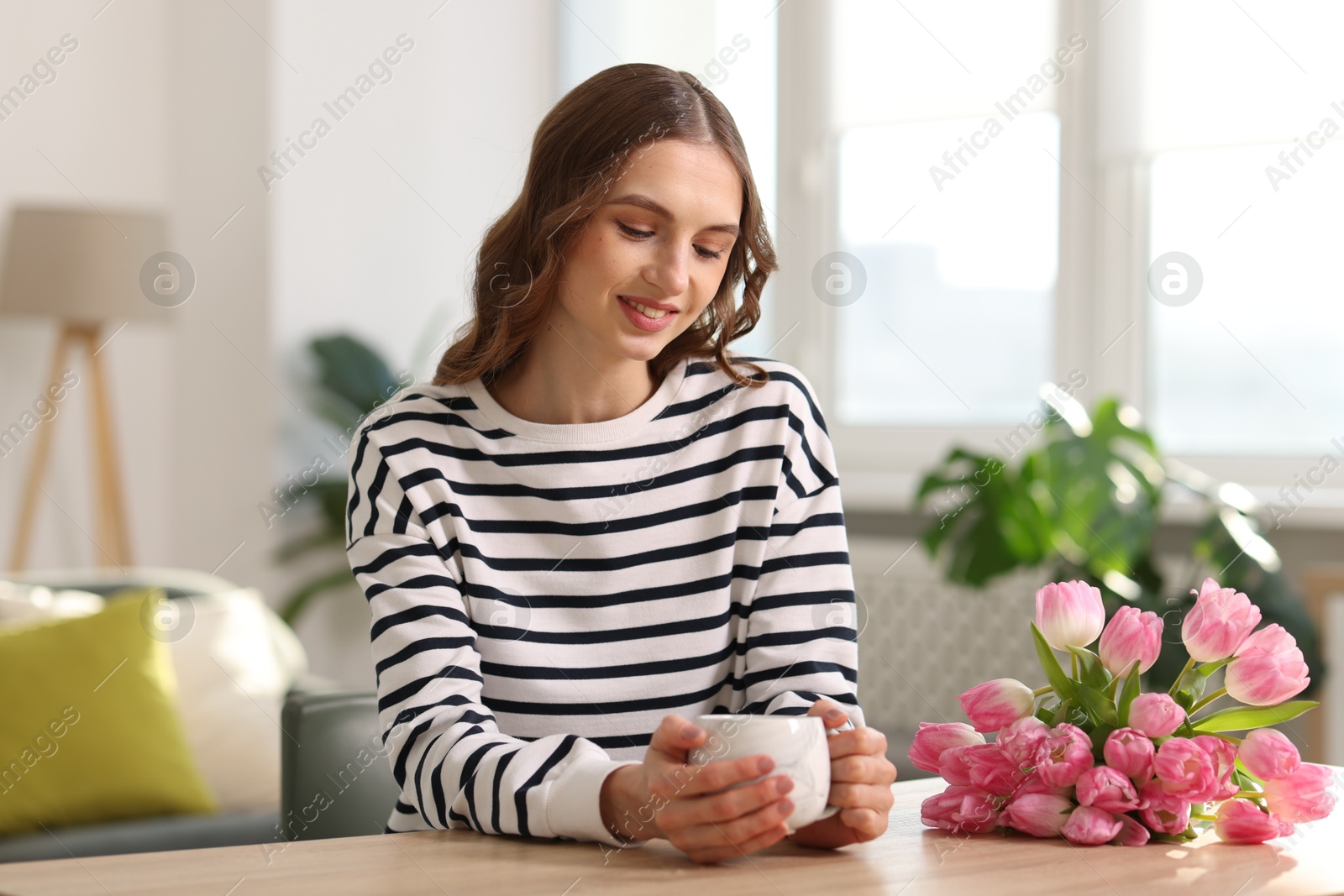 Photo of Smiling woman with bouquet of tulips drinking at table indoors