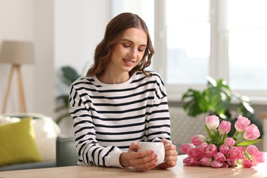 Photo of Smiling woman with bouquet of tulips drinking at table indoors