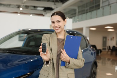 Photo of Happy saleswoman with clipboard and key near new blue car in salon, selective focus