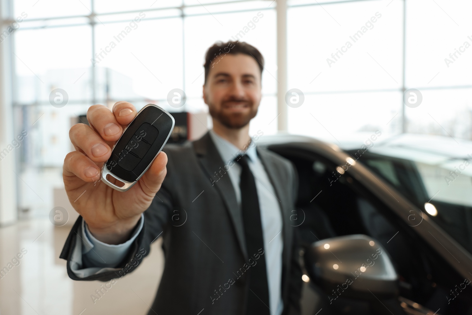 Photo of Happy salesman with key near new car in salon, selective focus