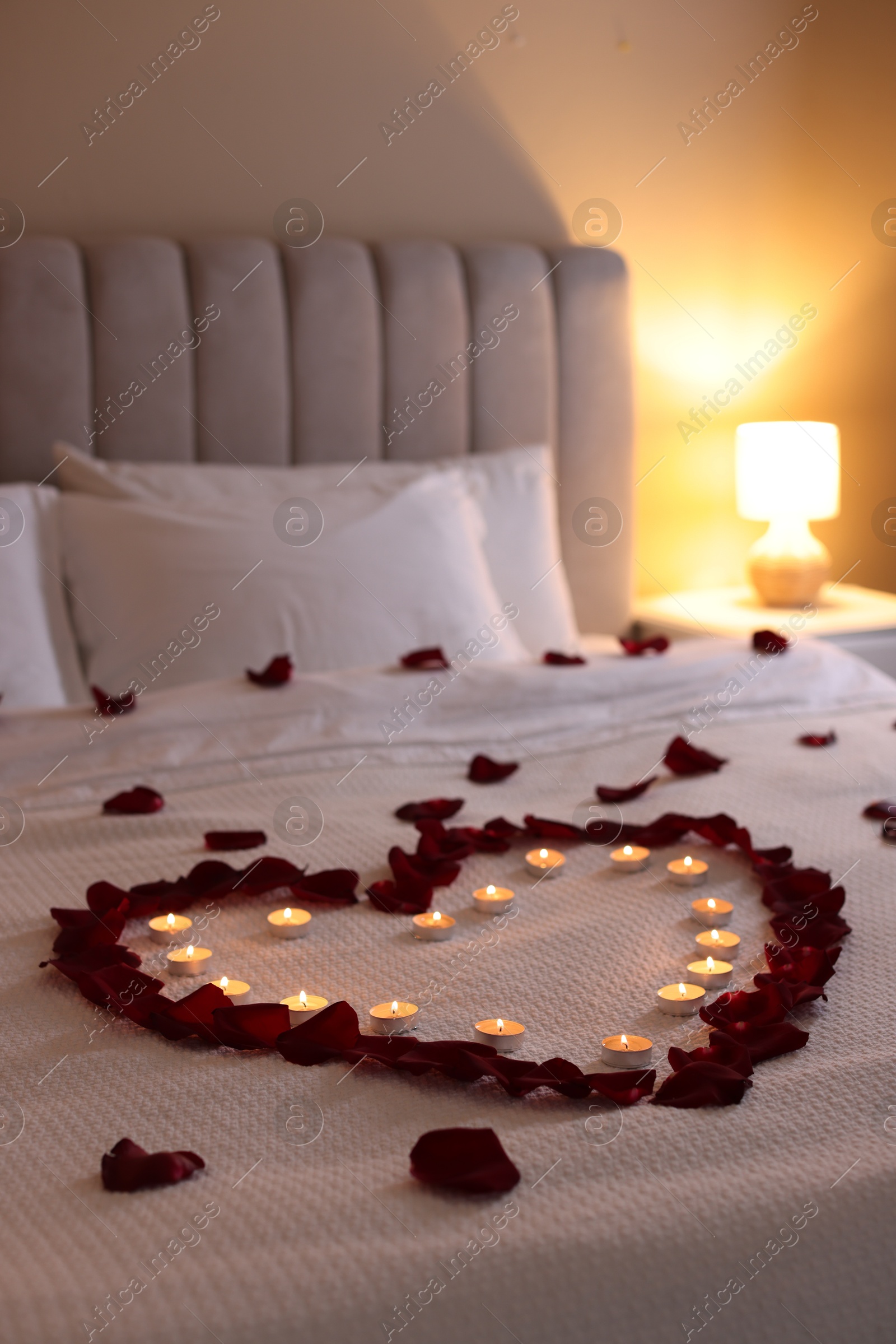 Photo of Heart made with beautiful rose petals and burning candles on bed indoors