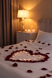 Photo of Heart made with beautiful rose petals and burning candles on bed indoors