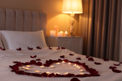 Photo of Heart made with beautiful rose petals and burning candles on bed indoors
