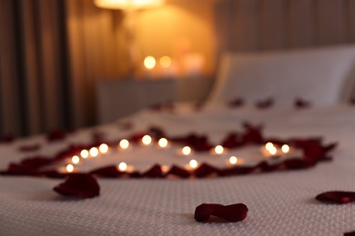 Photo of Heart made with beautiful rose petals and burning candles on bed indoors, selective focus