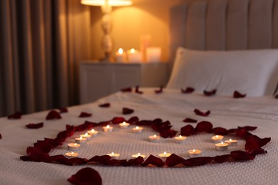 Photo of Heart made with beautiful rose petals and burning candles on bed indoors