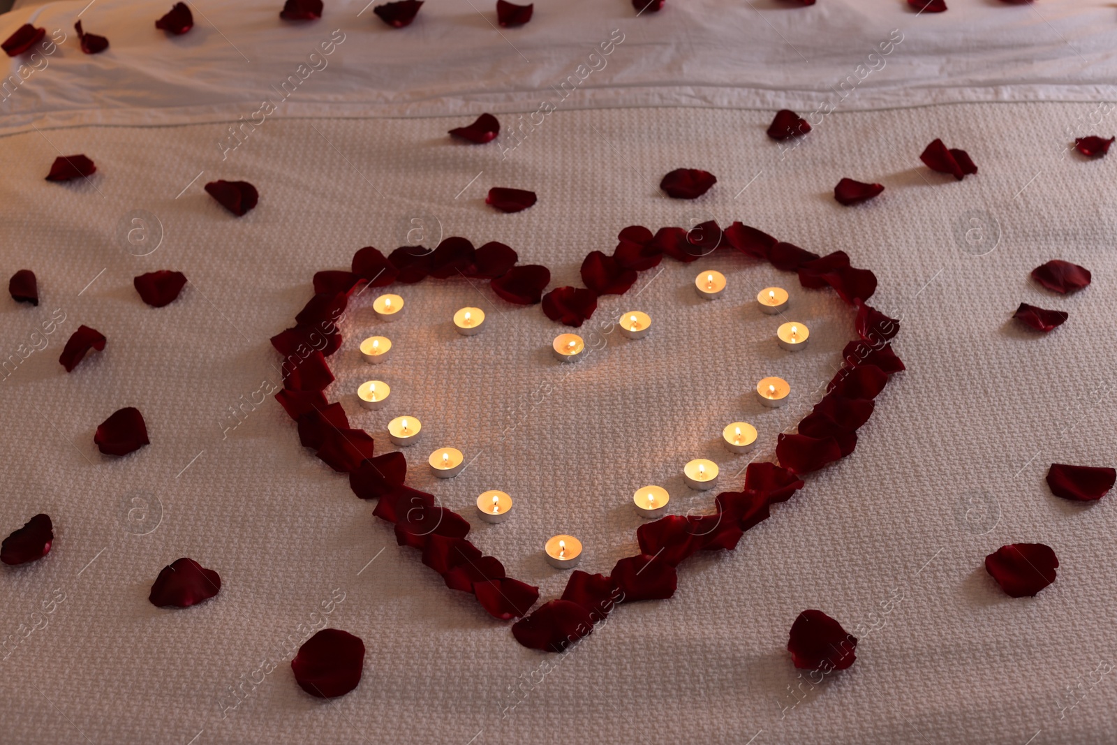 Photo of Heart made with beautiful rose petals and burning candles on bed