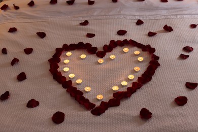 Photo of Heart made with beautiful rose petals and burning candles on bed