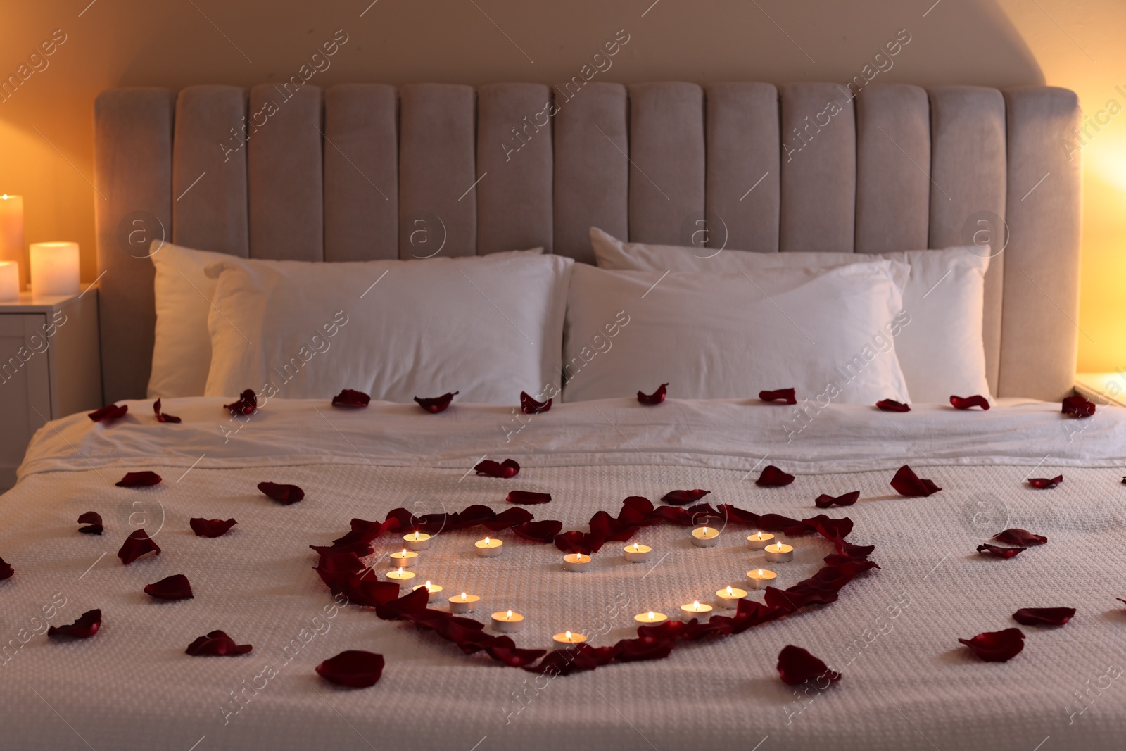 Photo of Heart made with beautiful rose petals and burning candles on bed indoors