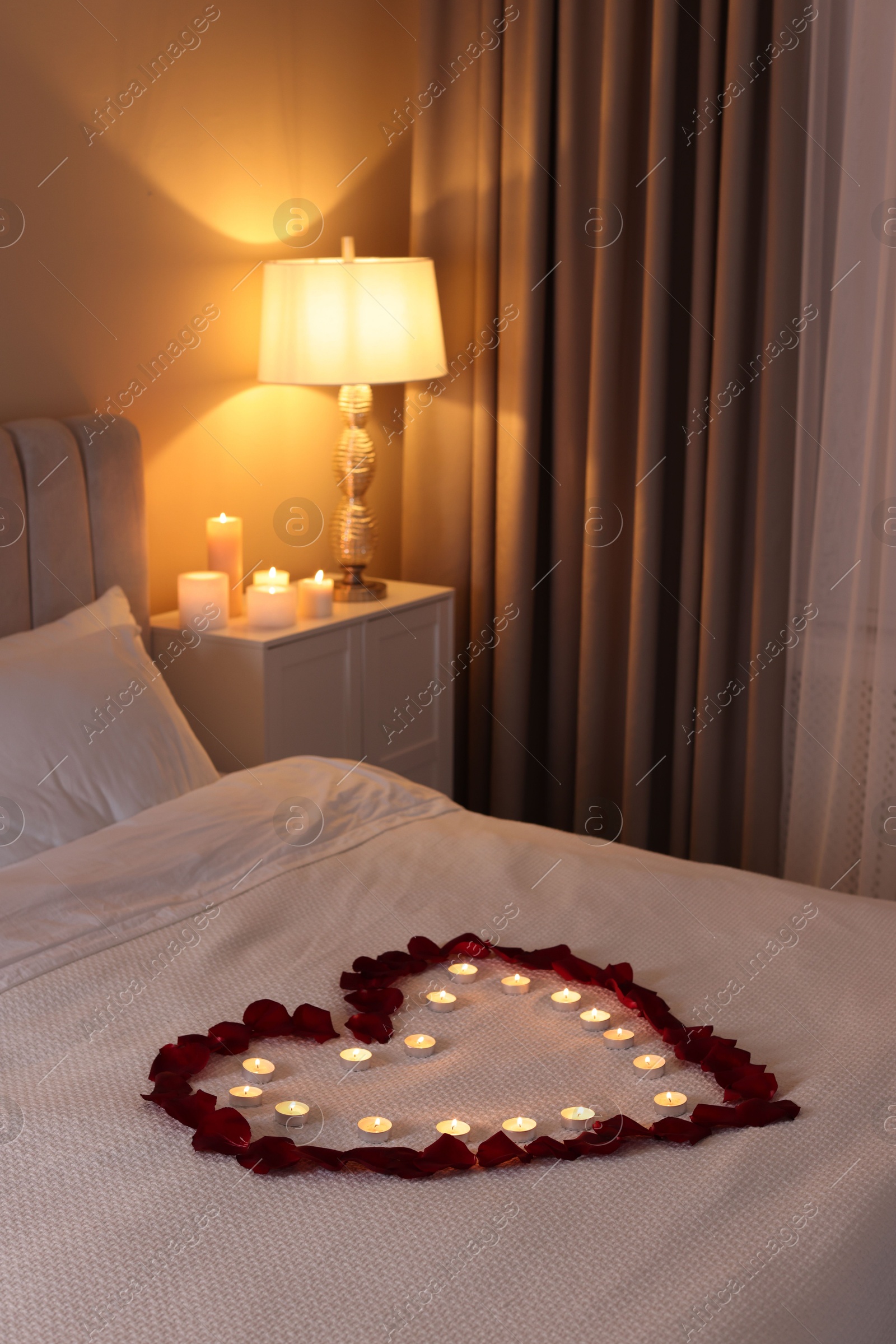 Photo of Heart made with beautiful rose petals and burning candles on bed indoors