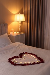 Photo of Heart made with beautiful rose petals and burning candles on bed indoors